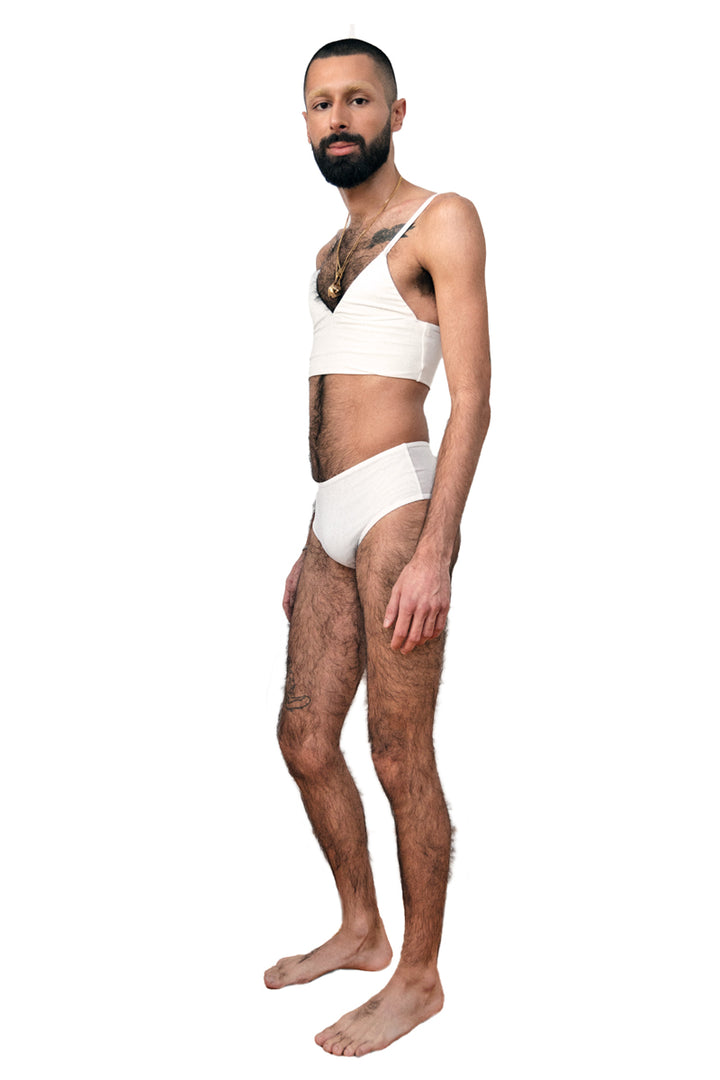Non-binary person wearing a white tucking underwear compression gaff with a cheeky boyshort cut with a white bamboo bra, photographed from the side.