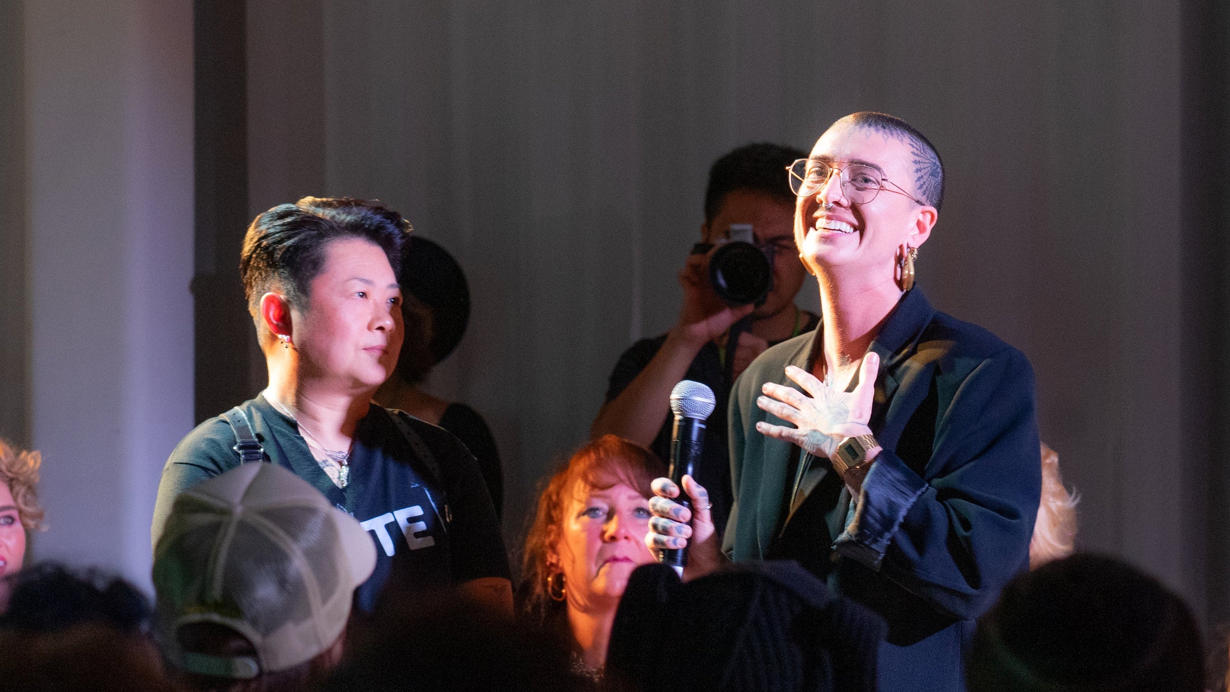 Rae Hill CEO or Origami Customs speaking into a handheld microphone on stage, surrounded by an audience and photographers in a dim indoor event setting.