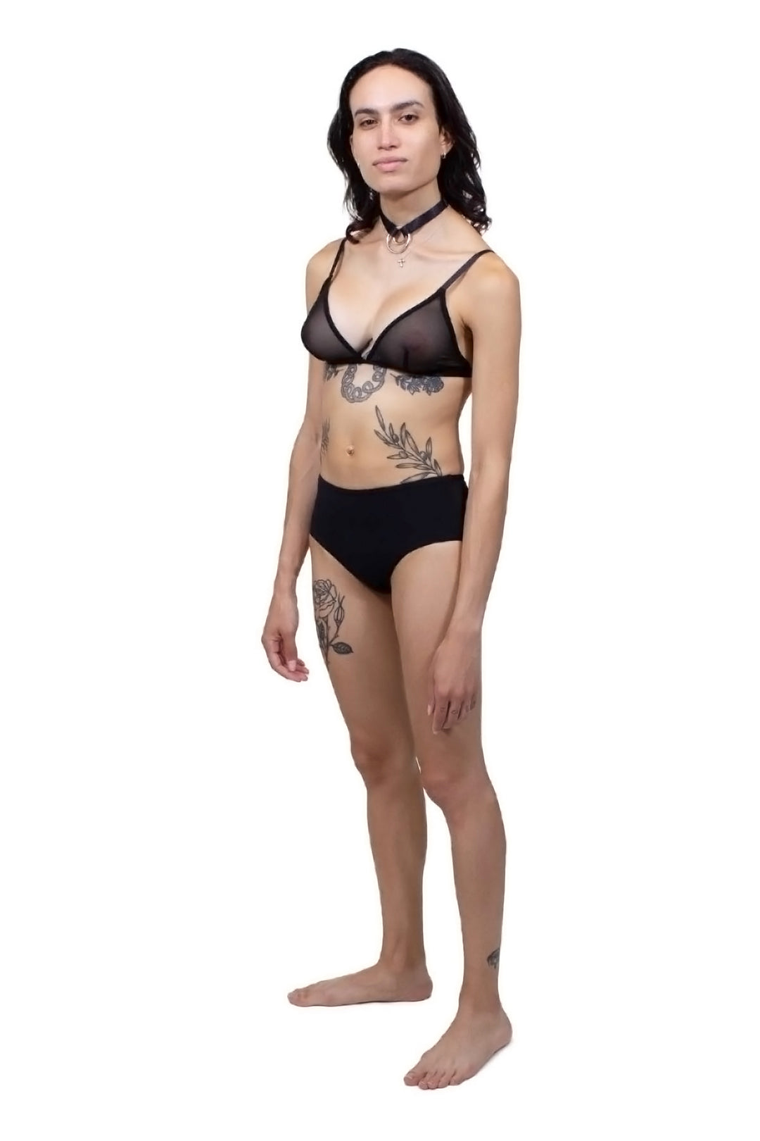 Transfemme person wearing a black tucking underwear compression gaff with a boyshort cut with a mesh bra, photographed from the left side.