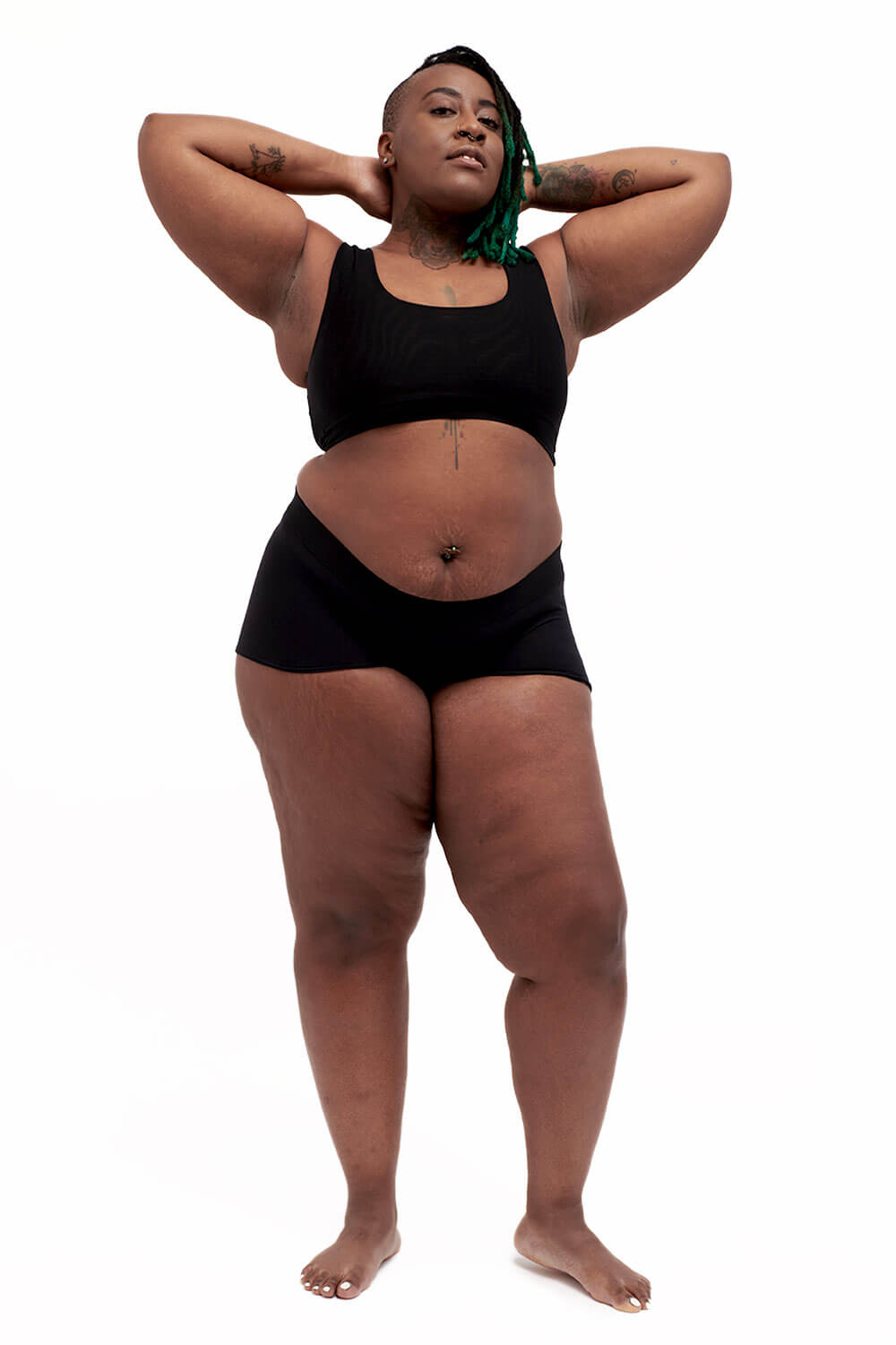 Plus-sized person wearing a black sports bra style chest binder made from breathable mesh, photographed from the front with arms up.