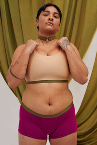 A Transgender non-binary model faces the camera. They are wearing the custom, sustainable collar in green with gold rings. They wear it with a gender- affirming custom chest binder. 