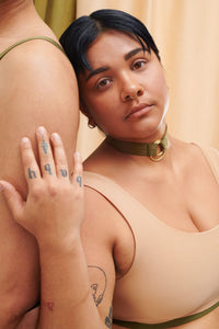 A Transgender non-binary model faces the camera. They are wearing the custom, sustainable collar in green with gold rings. They wear it with a gender- affirming custom chest binder. 