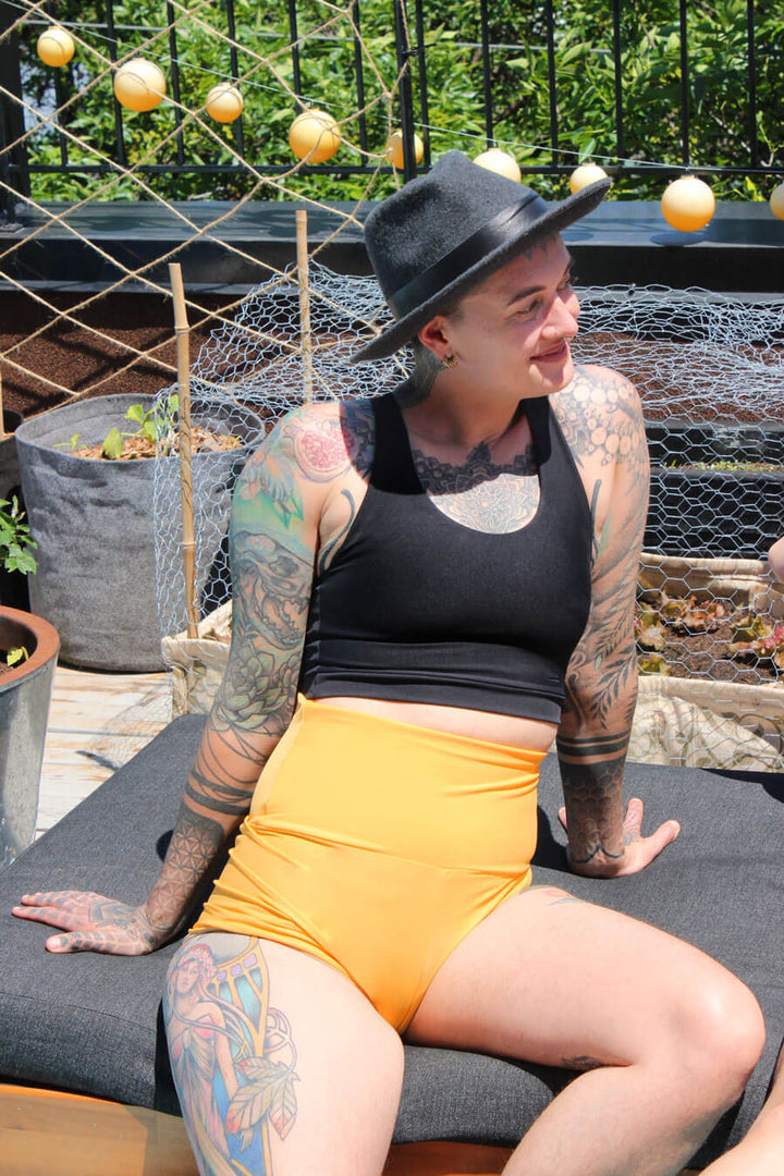 ENBY person wearing a black racerback soft chest binder made from lycra, and yellow gender affiming swim shorts, photographed from the front.