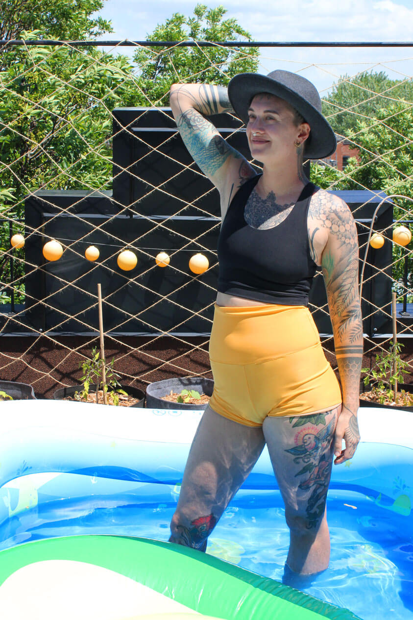 ENBY person wearing a black racerback soft chest binder made from lycra, and yellow gender affiming swim shorts, in a swimming pool and photographed from the front.